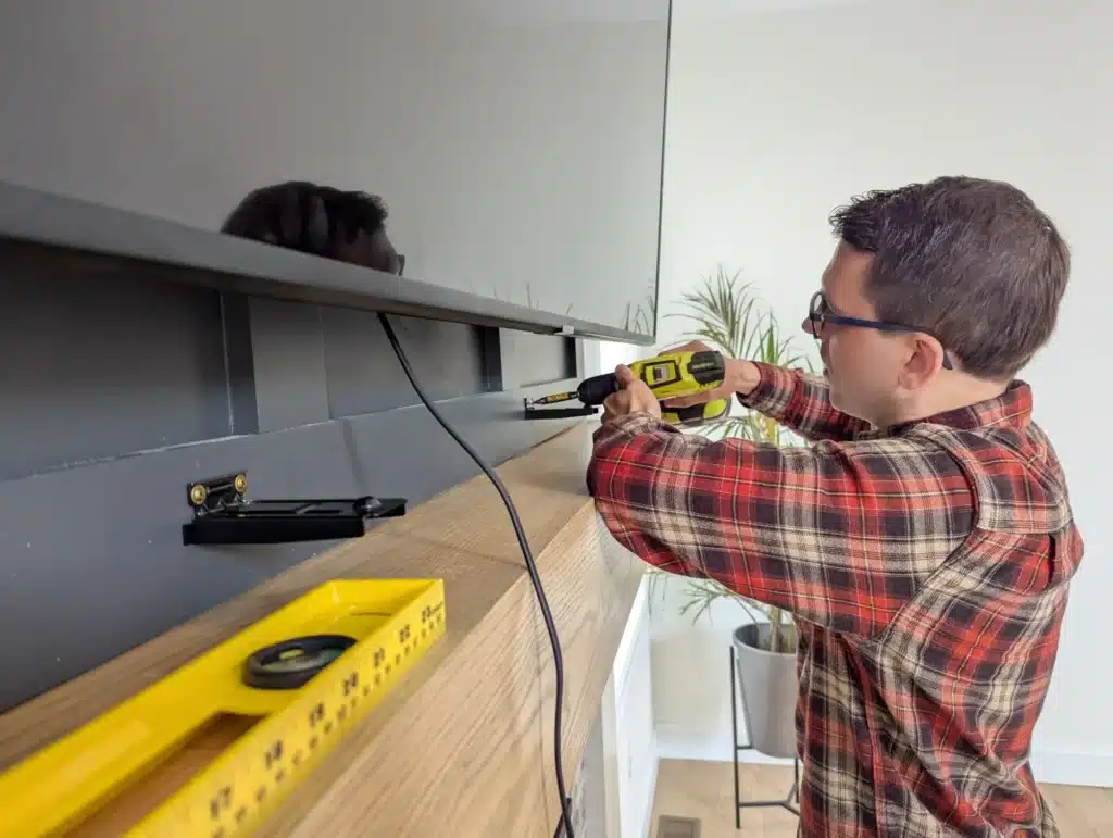 Horus Community Services technician drilling to install a floating media shelf beneath a wall-mounted TV in Trumansburg, NY