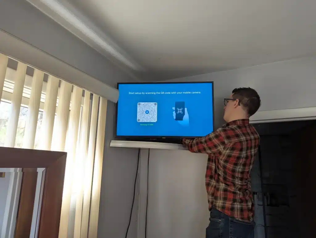Horus Community Services technician mounting a TV high on a living room wall near window blinds in Trumansburg, NY