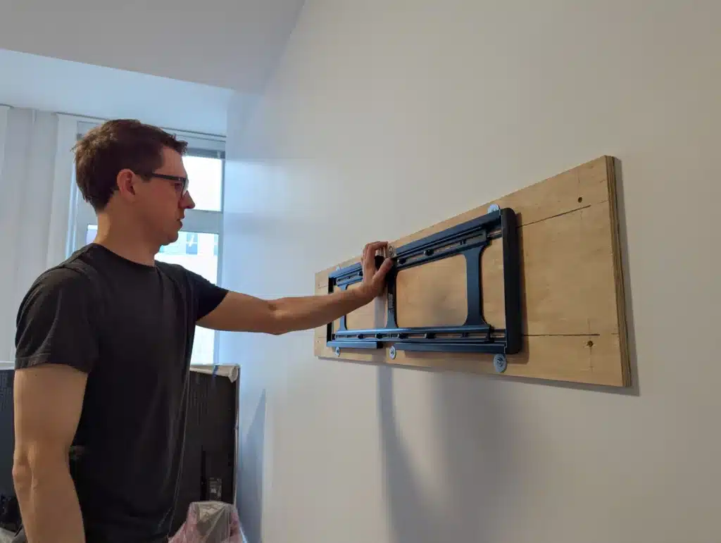 Horus Community Services technician fitting a Sanus TV mount plate onto a wood backer board in Trumansburg, NY