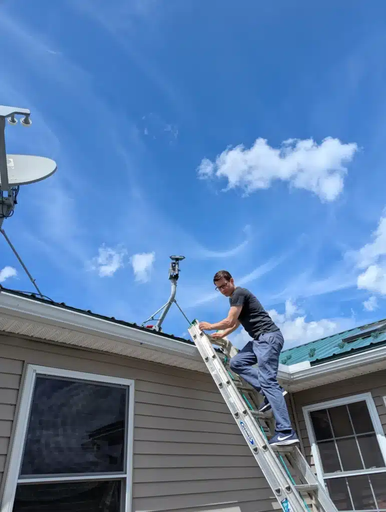 Horus Community Services technician installing a Starlink dish near the roofline from a ladder in Trumansburg, NY